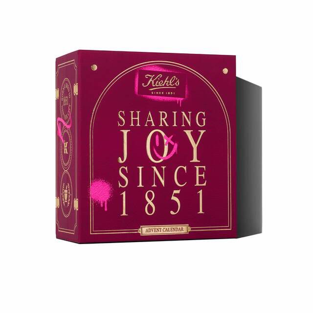 A deep red Kiehl’s Advent Calendar gift box with gold detailing and the text “Sharing Joy Since 1851” on the front. The box is shown angled to reveal the festive side panel artwork.