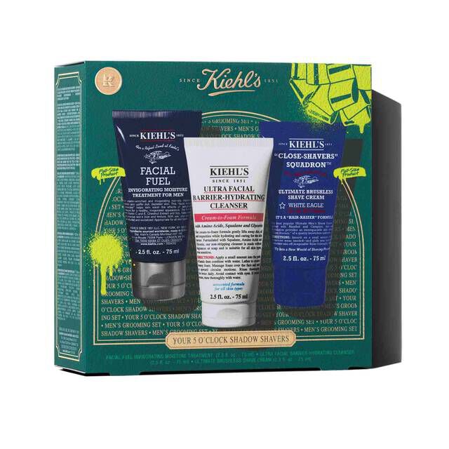 A graphic featuring a dark teal Kiehl’s gift box with gold accents. The box contains three products: a dark blue tube of "KIEHL’S FACIAL FUEL MOISTURIZER," a white tube of "KIEHL’S ULTRA FACIAL BARRIER-HYDRATING CLEANSER," and a dark blue tube of "KIEHL’S CLOSE-SHAVERS SQUADRON ULTIMATE BRUSHLESS SHAVE CREAM." The box is decorated with bright green graphic paint splatters and a bow design, and features the text "YOUR 5 O’CLOCK SHADOW SHAPERS" at the bottom.