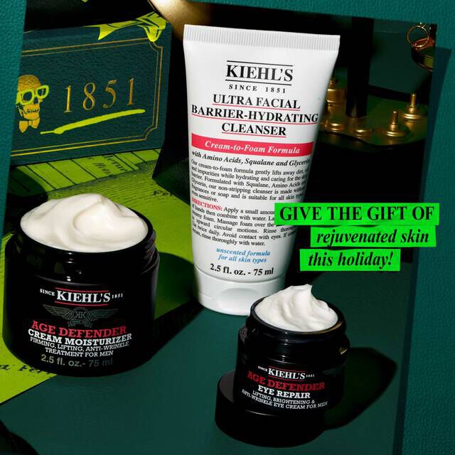 A festive graphic, set against a dark green background with a map pattern, features three Kiehl's products. A white tube of "KIEHL’S ULTRA FACIAL BARRIER-HYDRATING CLEANSER" is prominent. Below it is a black and red jar of "KIEHL’S AGE DEFENDER CREAM MOISTURIZER" and a smaller black and red jar of "KIEHL’S AGE DEFENDER EYE REPAIR," both with visible white cream. In the upper left background, a green gift box with "1851" and a skull design is visible, along with gold decorative elements. Bright green text in the middle right reads "GIVE THE GIFT OF rejuvenated skin this holiday!"