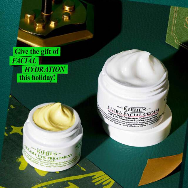 A graphic displaying a white jar of "KIEHL'S CREAMY EYE TREATMENT with avocado" showing a yellow-green product swirl and a larger white jar of "KIEHL'S ULTRA FACIAL CREAM" with a white product swirl. They are set on a dark green surface with gold accents, including a decorative gold object in the upper left. Green text in the upper left corner reads "Give the gift of FACIAL HYDRATION this holiday!".